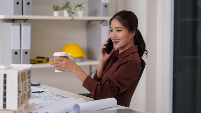 Asian woman architect professional consults building project development design plans via phone hold coffee cup modern office environment construction management
