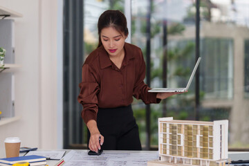 Asian adult woman architect designs architectural blueprints laptop building model modern office professional work planning future