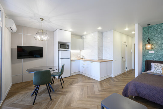 bright, modern studio apartment featuring a small kitchen, dining area with a TV, and a cozy bedroom space. The design is clean and efficient, with herringbone floors and contemporary lighting