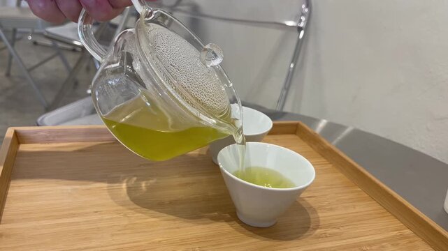 Pouring fresh green tea from glass teapot into white ceramic cup on wooden tray, symbolizing relaxation, mindfulness, Asian tea culture, healthy lifestyle, and peaceful refreshment