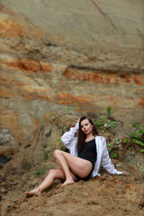 Young beautiful long legged brunette woman in a black swimsuit and white shirt sitting against the background of a sandy cliff