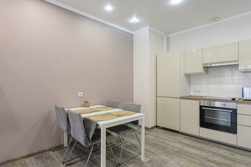 well-lit kitchen with a dining area. The kitchen features light beige cabinets and appliances. A white table with grey chairs sits to the side. Wooden floor