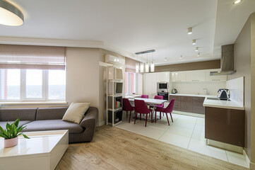 bright, modern open-concept living and kitchen area. Features a gray sofa, dining table with pink chairs, and a contemporary kitchen with white and brown cabinets