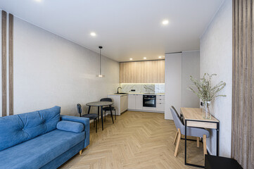 modern apartment interior featuring a blue sofa, dining table, kitchen with wooden cabinets, and herringbone floors. Stylish lighting and decor add to the contemporary aesthetic