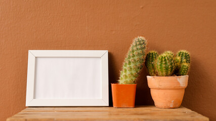 A cozy arrangement of two cacti in terracotta pots sits on a wooden table, with a blank white frame leaning against a warm brown wall, creating a simple, minimalist decor