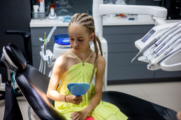 Young girl in a dental clinic preparing for treatment while holding a mirror and sitting in a comfortable dental chair