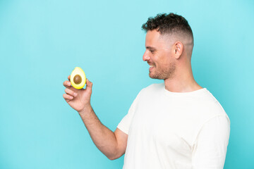 Young caucasian man holding an avocado isolated on blue background with happy expression