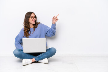 Young caucasian woman with a laptop sitting on the floor isolated on white background pointing finger to the side and presenting a product