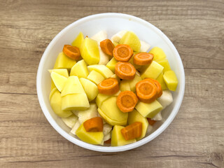 Chopped potatoes, carrots, and root vegetables in a white bowl on wooden table, prepared for cooking soup or stew.