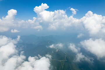 Jiugong Mountain in Xianning City, China, shows a vast landscape with lush green mountains. White, fluffy clouds float through the clear blue sky, partially obscuring the distant mountain ranges. S