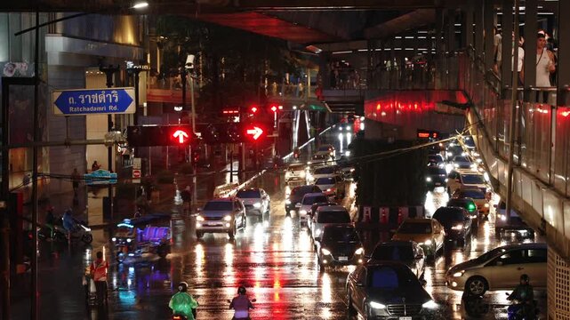 Rainy night traffic with reflections on wet road and people crossing by skywalk at Siam station