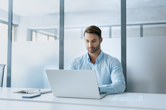 Young professional specialist latin hispanic business man working on laptop pc sitting at desk in modern office space. European businessman entrepreneur using computer technology app for work online