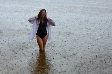 Beautiful young brunette woman in black swimsuit and white shirt standing in water of the sea near sandy shore in rainy weather