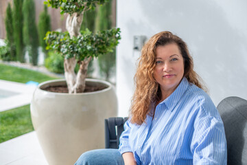 Middle-aged woman with curly hair, wearing blue shirt, sitting gracefully in a serene outdoor environment, exuding beauty, joy, and elegance in her life journey