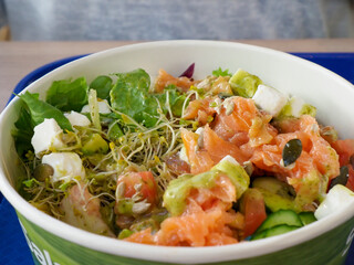 A close-up view of a fresh salmon salad with greens, sprouts, and vegetables in a white paper bowl on a blue tray.