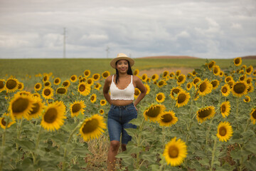Fototapeta premium Young, attractive, Latina brunette woman wearing a white crochet top, jeans, and a hat, smiling and happy as she walks through a field of sunflowers. Concept: nature, agriculture, countryside.
