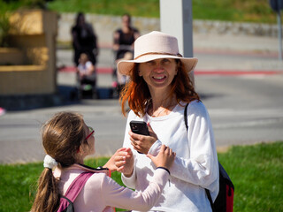 A woman in a summer dress and hat showing something on her phone to a young girl.