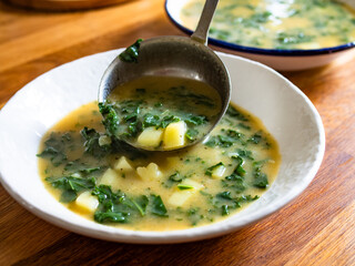 Pouring Portuguese green soup - Caldo Verde. Potato and kale soup on wooden table	
