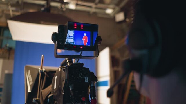 Headset cameraman monitors camera during studio shoot, recording female presenter in red dress against blue screen, teleprompter visible, soft light shining, control gear filling production area