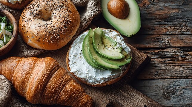 Close up of a fresh bagel topped with creamy cheese and ripe avocado slices, served alongside croissant and avocado halves on rustic wooden background. Perfect image for National Sandwich Day, breakfa