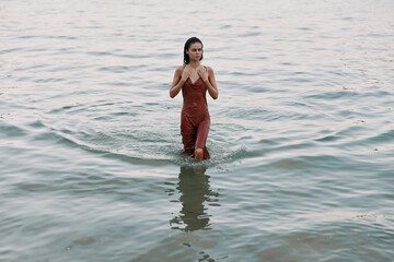 A woman stands knee-deep in calm water, wearing a rust red dress, creating gentle ripples as waves lap around her, capturing a serene intimate seaside moment.
