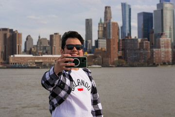 Smiling young Hispanic man taking selfie with smartphone during boat ride on Hudson River with iconic Manhattan skyline in background enjoying travel, lifestyle and urban tourism in New York City