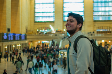 Smiling young Hispanic backpacker inside Grand Central Terminal in New York City surrounded by commuters and tourists enjoying lifestyle, architecture and urban tourism in iconic station