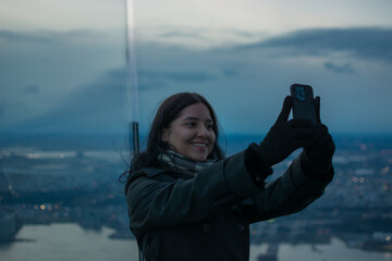 Smiling young Hispanic woman taking selfie with smartphone at sunset on a skyscraper rooftop in New York City enjoying authentic urban lifestyle and travel experience