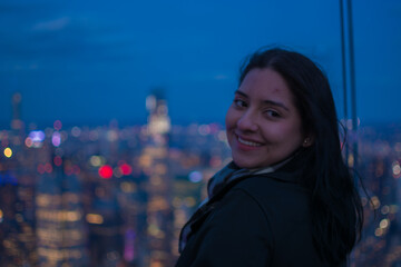 Smiling young Hispanic woman looking back on a New York City skyscraper rooftop at night with blurred city lights in the background enjoying authentic travel lifestyle experience