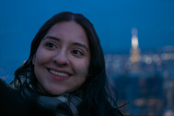 Close up of smiling young Hispanic woman at night on New York City skyscraper rooftop with glowing Empire State Building in the background enjoying authentic urban lifestyle and tourism