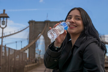 Smiling young Hispanic woman drinking bottled water while enjoying a sunny day on the Brooklyn Bridge in New York City urban lifestyle and tourism scene