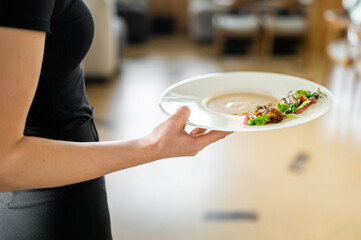 Waiter holding a white plate with gourmet dish, elegantly plated with sauce, herbs, and vegetables. Fine dining presentation in warm indoor restaurant setting.