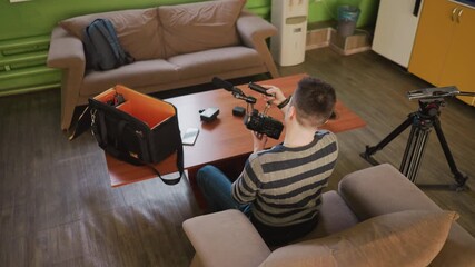 Young man sets up camera equipment, removing microphone and camera gear in backpack. Casual home environment with sofa and wooden table. Professional filming preparations in cozy space
