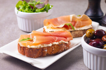 Two bruschettas with prosciutto, ricotta cheese and rucola on ciabatta bread. Olive and spices  decorated.