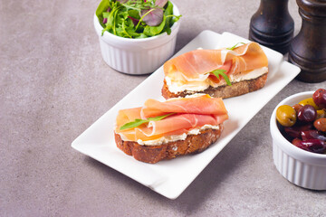 Two bruschettas with prosciutto, ricotta cheese and rucola on ciabatta bread. Olive and spices  decorated.