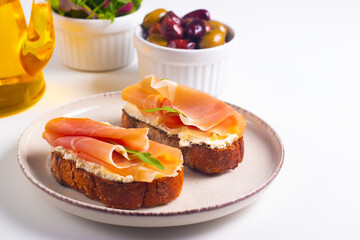 Two bruschettas with prosciutto, ricotta cheese and rucola on ciabatta bread. Olive and spices  decorated.