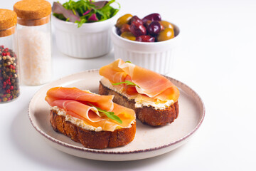 Two bruschettas with prosciutto, ricotta cheese and rucola on ciabatta bread. Olive and spices  decorated.