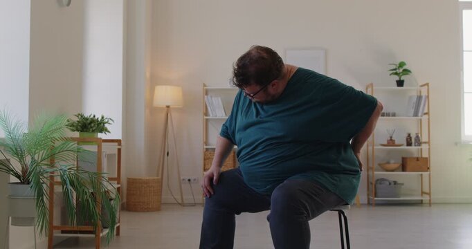 Fat overweight obese man presses his lower back while sitting on a chair at home, expressing pain and discomfort linked to poor posture or muscle strain. Man with backpain holds lowerback - Powered by Adobe