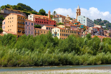 A view of a beautiful Italian old town Vintimille rising up into the hillside towards mountains in the distance. There are painted houses and church spire and wooded hills. © kovalenkovpetr