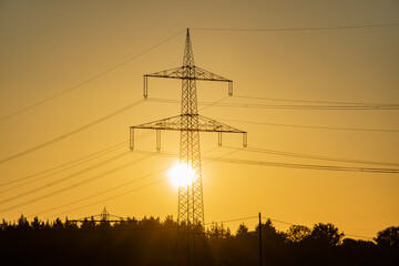 Transmission tower or high-voltage electricity pylon with sun shining through steel frame at sunset. Symbol of energy and power generation.