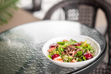 Fresh salad with leafy greens, cherry tomatoes, apple slices, grilled beef, and seeds in a white bowl on a glass table outdoors. Healthy meal in natural light, patio setting