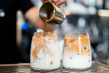 Barista pouring espresso into iced milk in glass, creating layered iced coffee. Close-up in café setting, showcasing beverage preparation and refreshing drink concept.