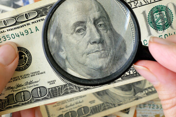 hand with magnifying glass checking 100 USA dollar banknote on money background, exchange money