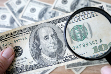 hand with magnifying glass checking 100 USA dollar banknote on money background, exchange money