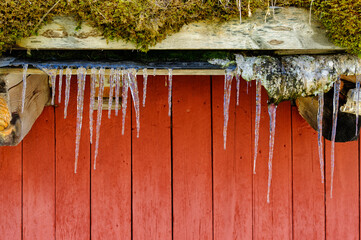 Icicles form under a weathered wooden eave, contrasting against a vibrant red wall, showcasing winter's chill and beauty.