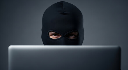 Hacker in black mask using laptop for cybercrime or anonymous online activity