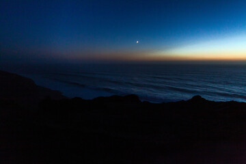 A serene twilight coastal scene with a crescent moon glowing above the calm ocean,