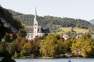 Fototapeta premium Scenic view of St. Martin's Church in Bled, Slovenia, with its tall spire rising above trees and hills, and a traditional pletna boat floating on Lake Bled.