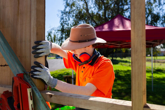 Young trainee apprentice worker with gloves holding wood to frame of building during traineeship