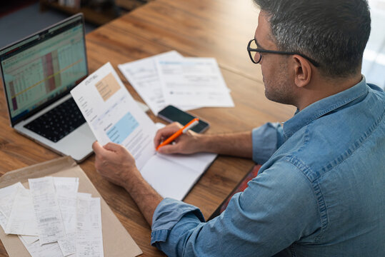 Mature man with glasses holds utility bill writing in paper notebook at laptop, male accountant calculates expenditures for company office service works, financial reports